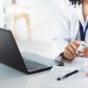 General practitioner holding a medicine bottle in front of a laptop on a table.
