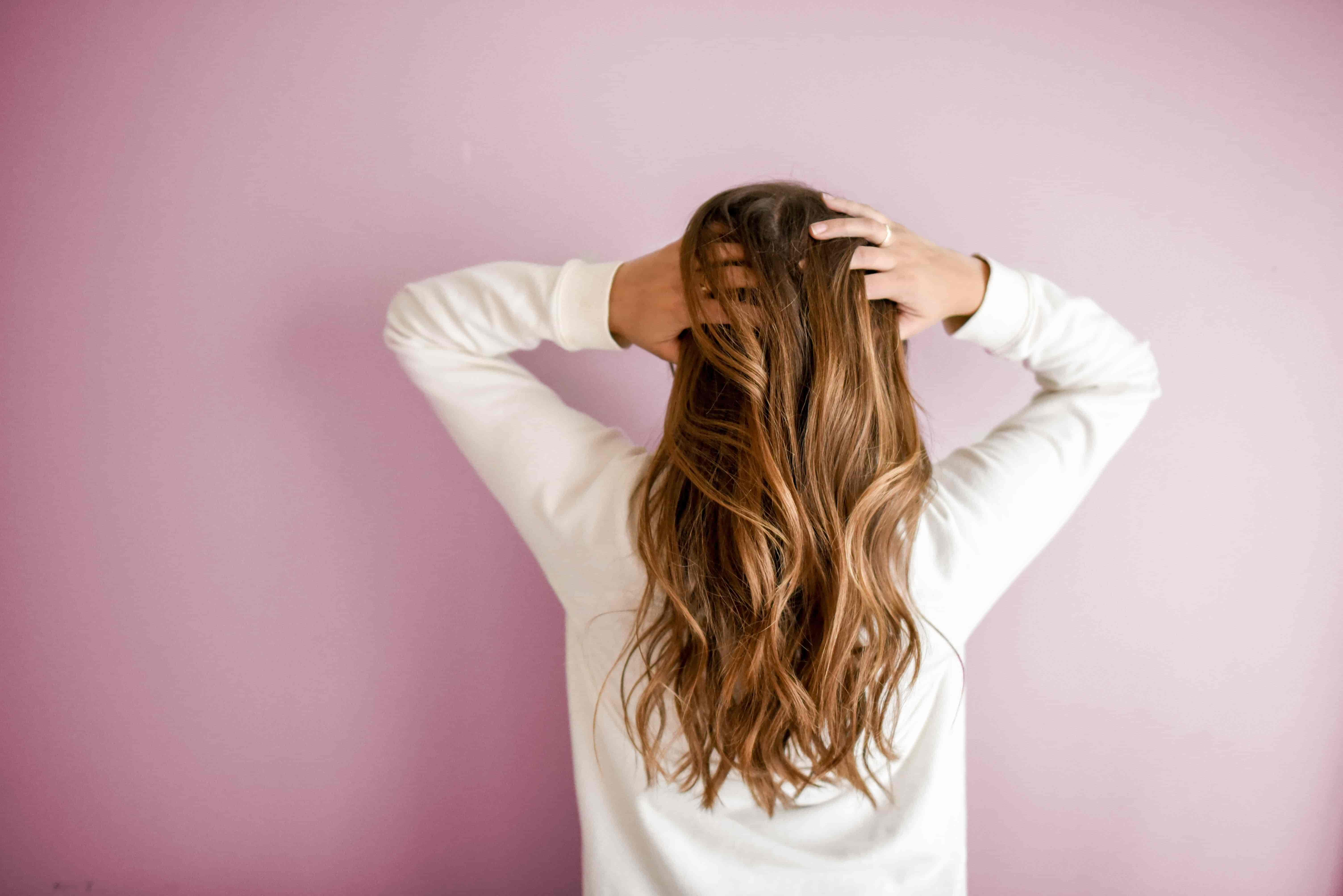 A woman in a cream jumper with long wavy brown hair, with her back towards the camera with both hands on her head on a pink background