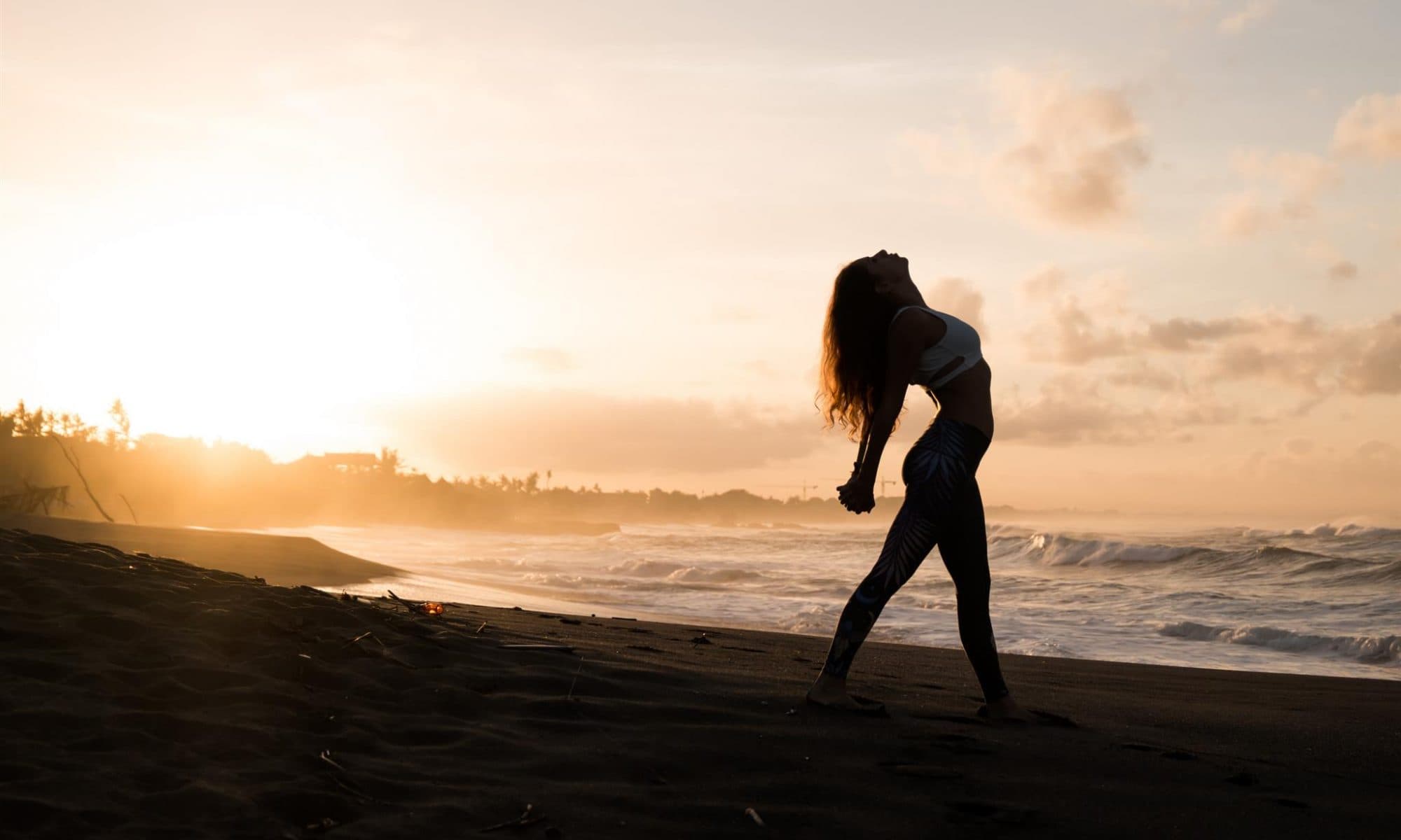Woman performing yoga on the beach with sun setting in the background