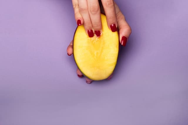 A woman with red nails putting her fingers into a half-cut mango