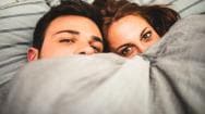 A man and woman in bed hiding behind a duvet